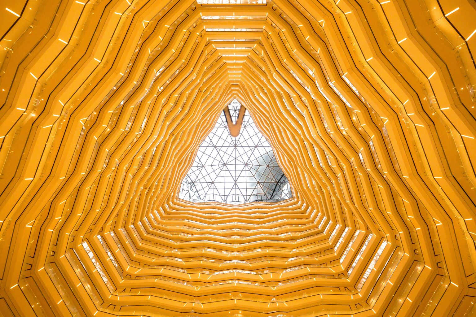 A triangular architectural ceiling design with layered yellow structures resembling honeycomb patterns, leading up to a glass skylight at the apex.