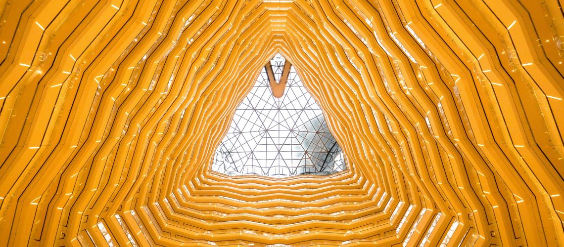 A triangular architectural ceiling design with layered yellow structures resembling honeycomb patterns, leading up to a glass skylight at the apex.